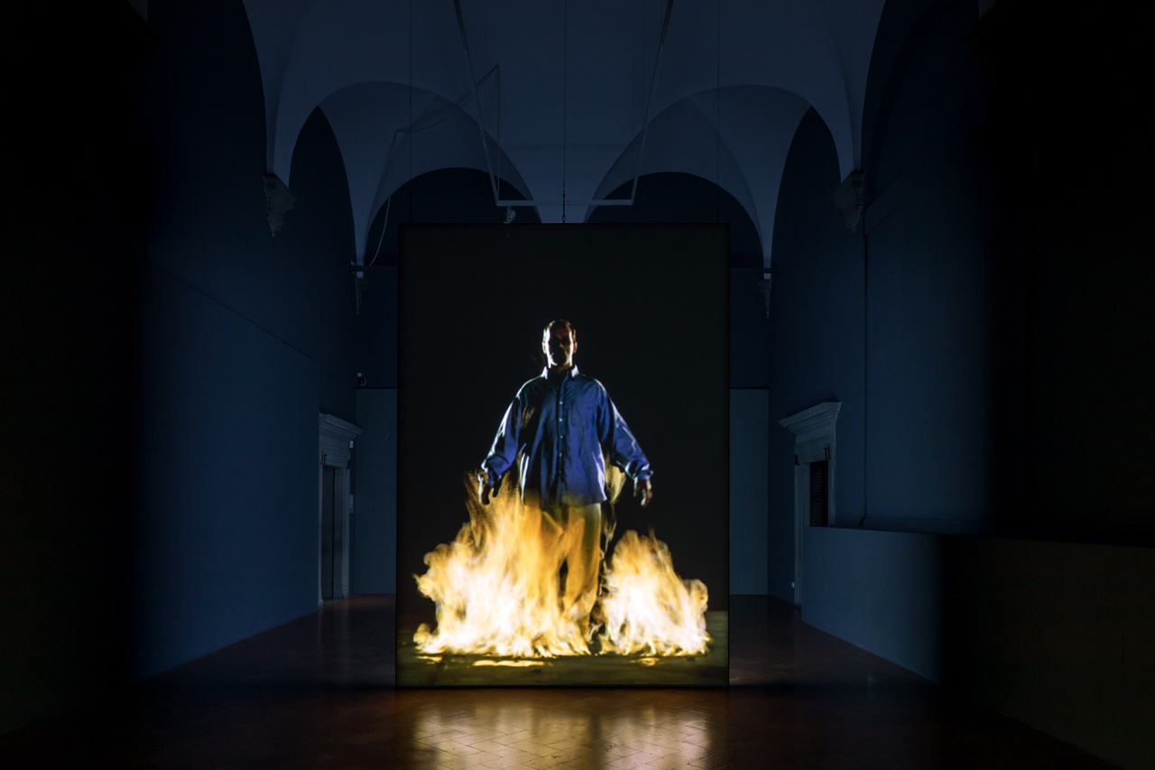 Still uit videokunstwerk The Crossing, van Bill Viola. Een man loopt op de kijker af. Een vuur laait op onder zijn voeten.